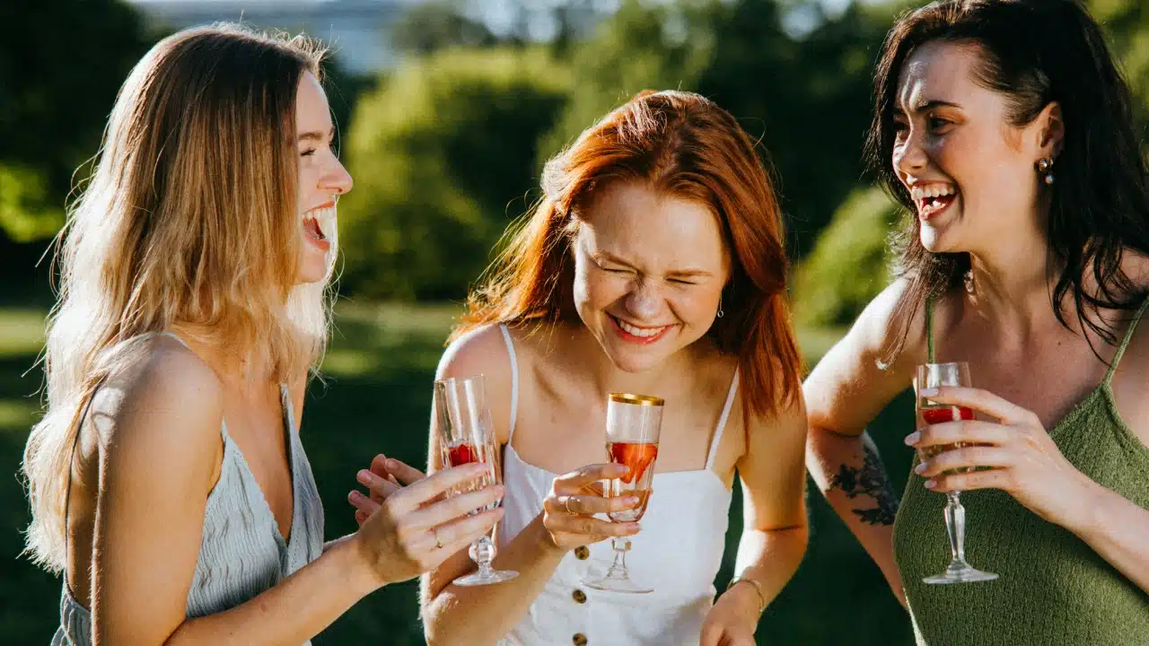 Three women stand outdoors in sunlight, laughing and holding glasses containing liquid and fruit.