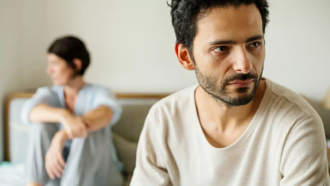 A man with dark hair looks away while a woman sits blurred in the background.