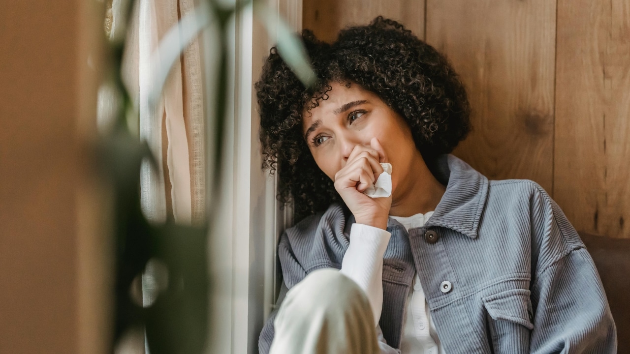 A woman with curly hair sits by a window holding a tissue to her face.