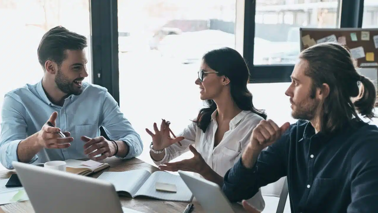 A woman wearing glasses gestures while talking with two men sitting at a wooden table.