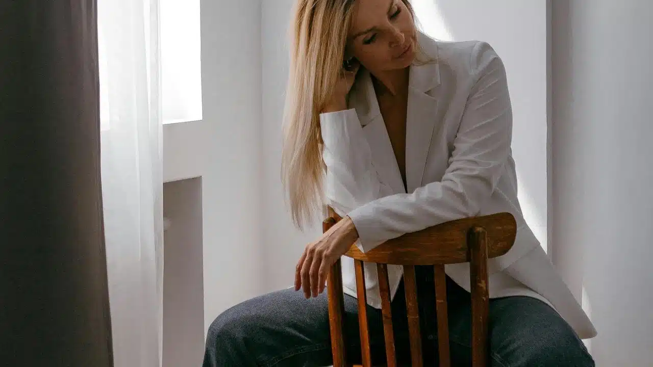 A woman in a white blazer sits on a chair, looking down thoughtfully.