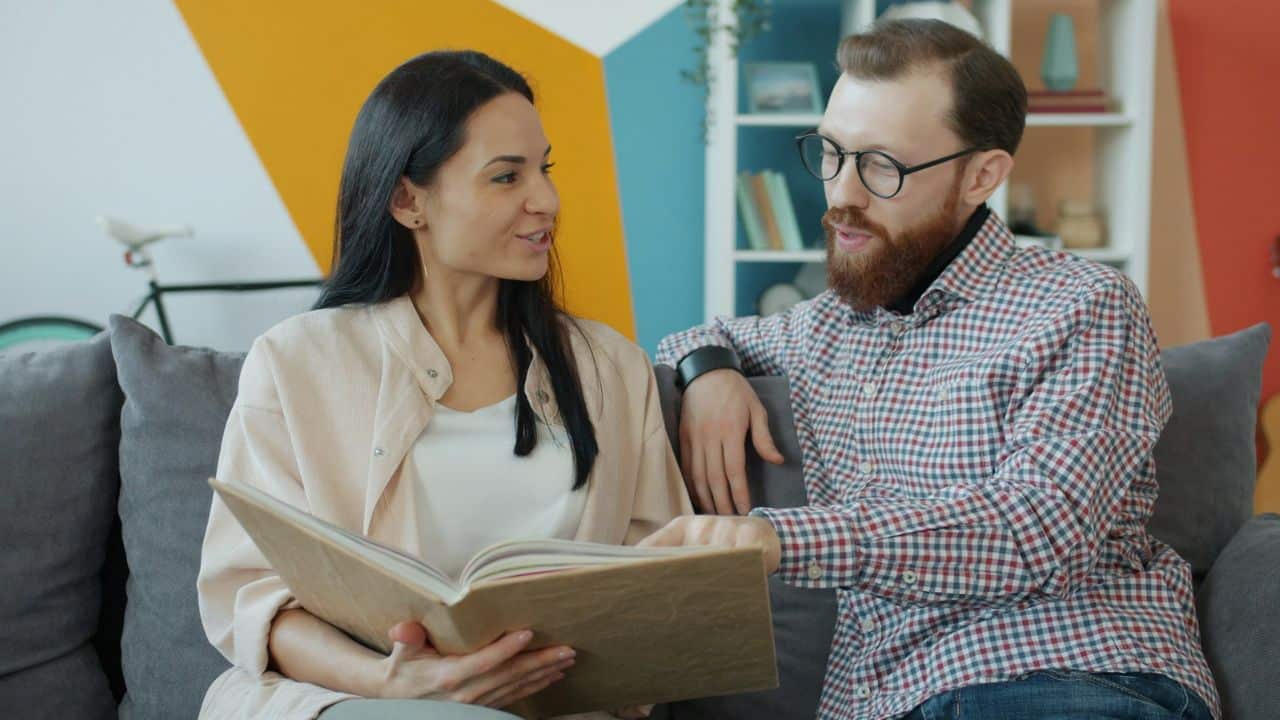 A woman and a man sit together on a sofa, looking at an open book.