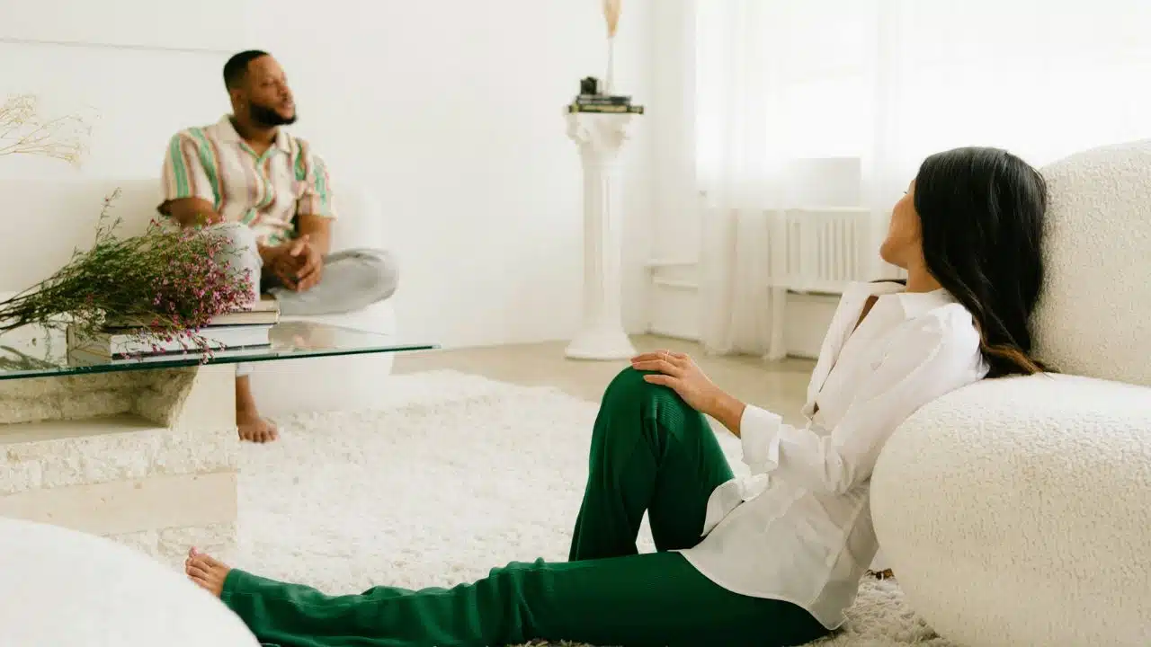 A man and woman sit apart in a bright, white room with minimalist furniture.