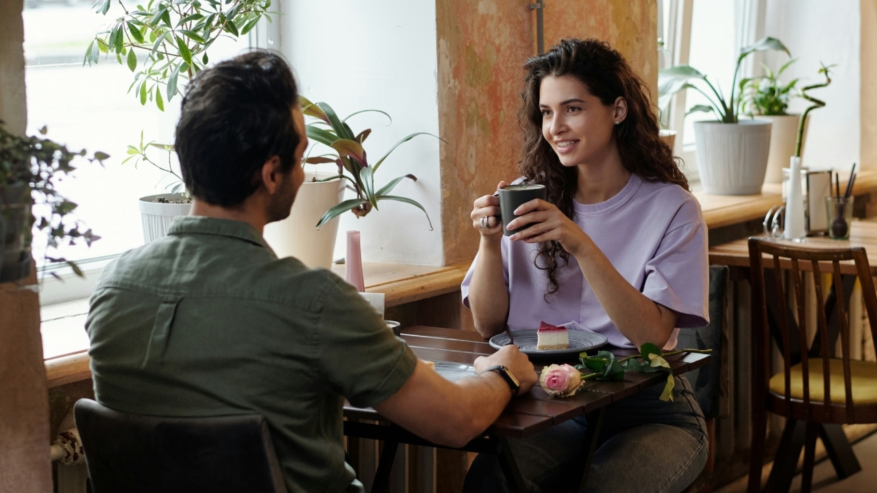 A man and woman sit at a small cafe table near a window with many potted plants.