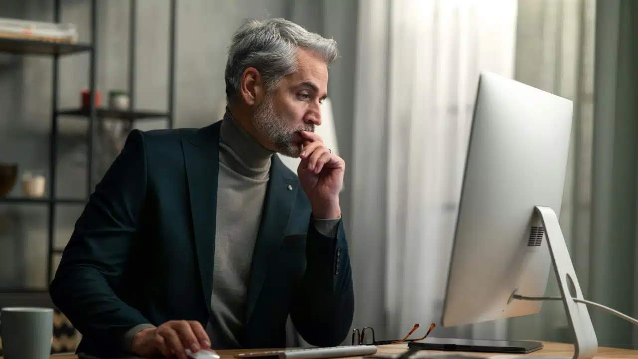 A gray-haired man in a dark blazer looks intently at a large desktop computer monitor.