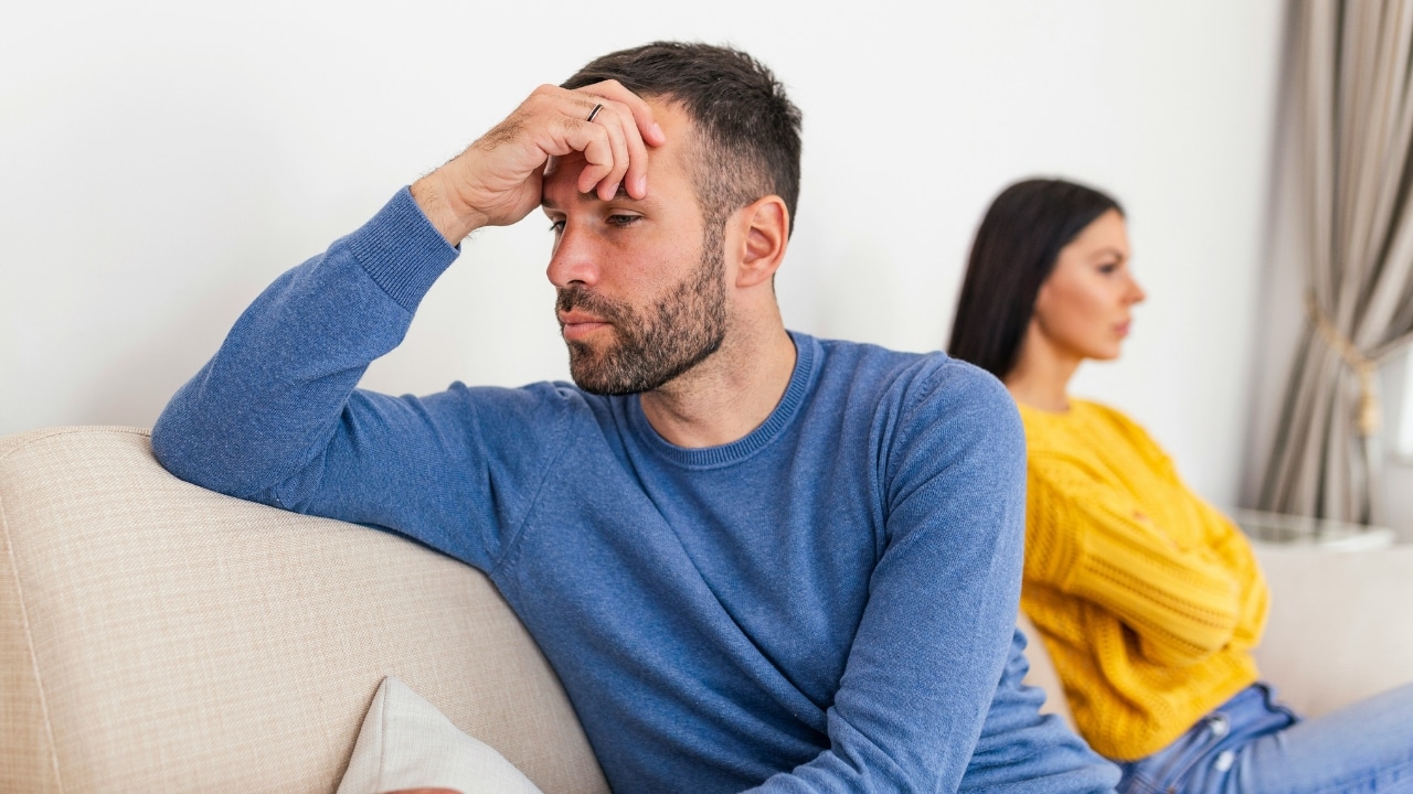 A man sits on a couch holding his head while a woman sits behind him.