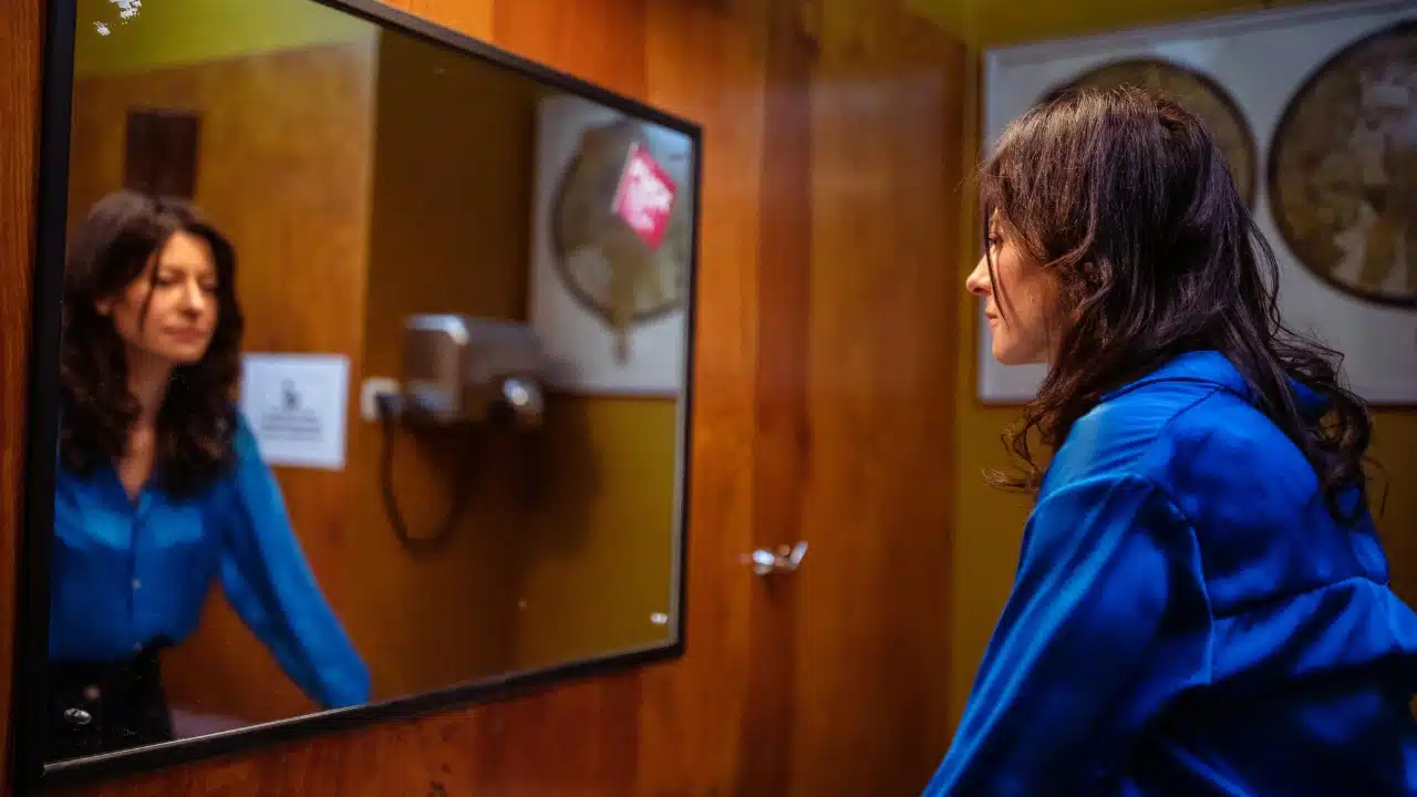 A woman in a blue shirt looks at her reflection in a bathroom mirror.
