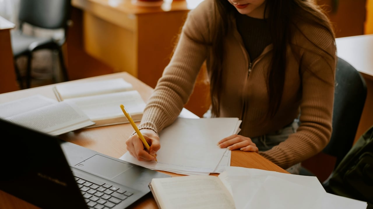 A person in a tan sweater writes on paper at a desk with a laptop.