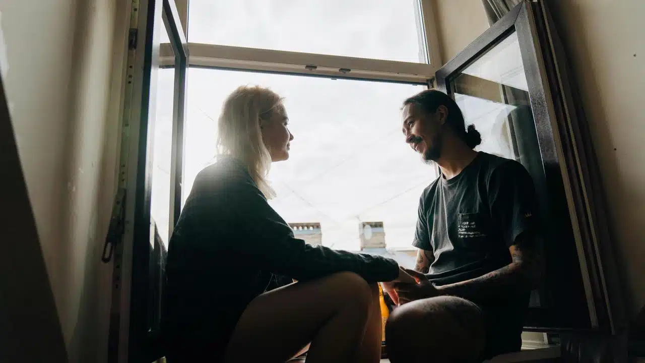 A couple sitting by an open window, talking and holding hands.