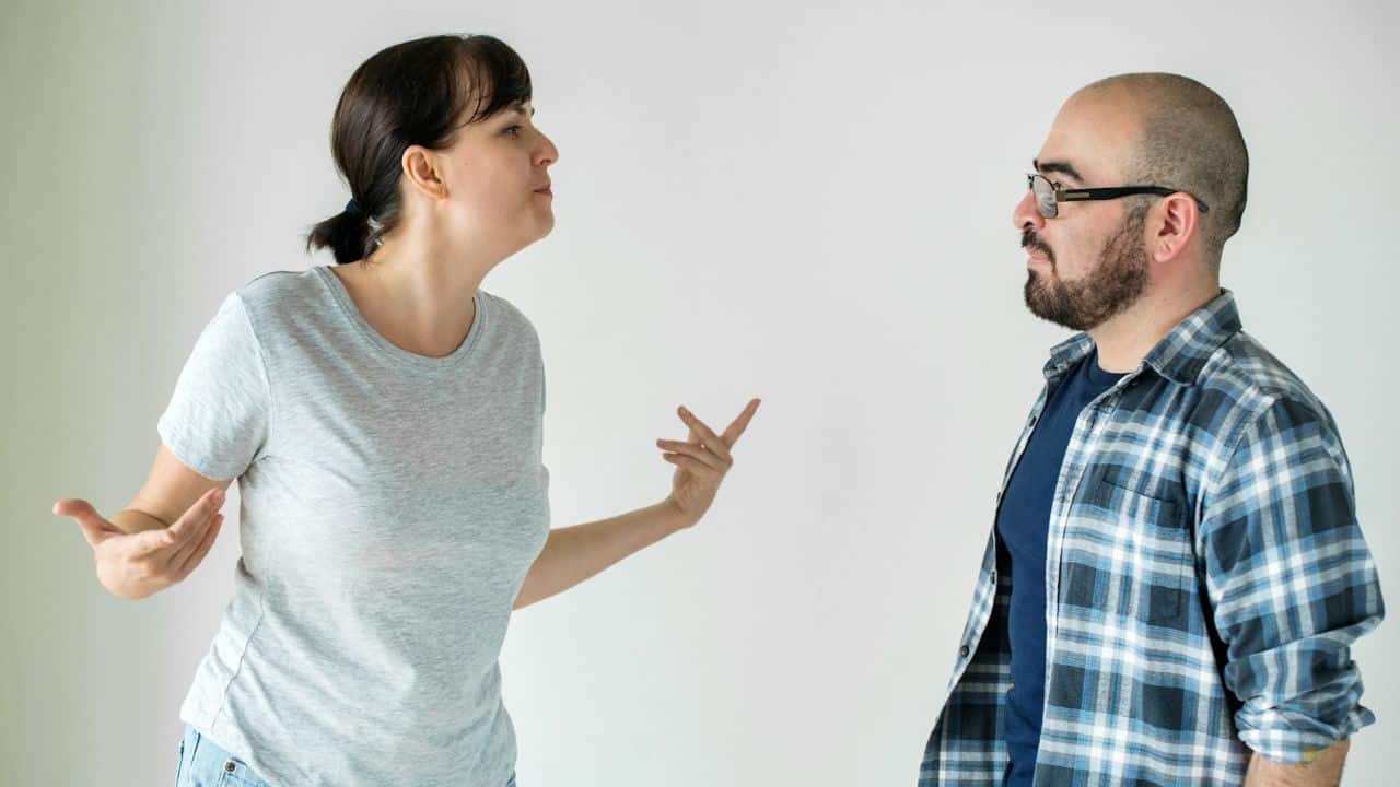 A woman gestures with both hands while standing across from a man wearing glasses.