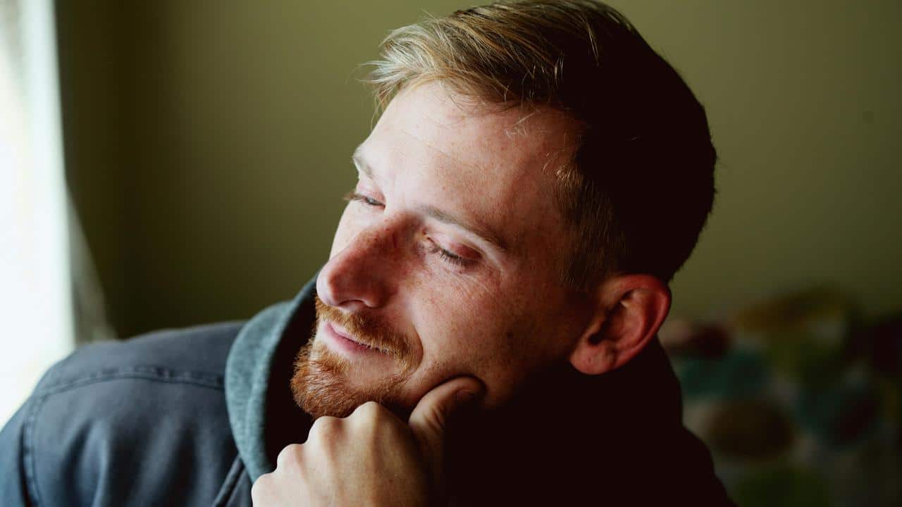 A man with a thoughtful smile resting his chin on his hand by a window.