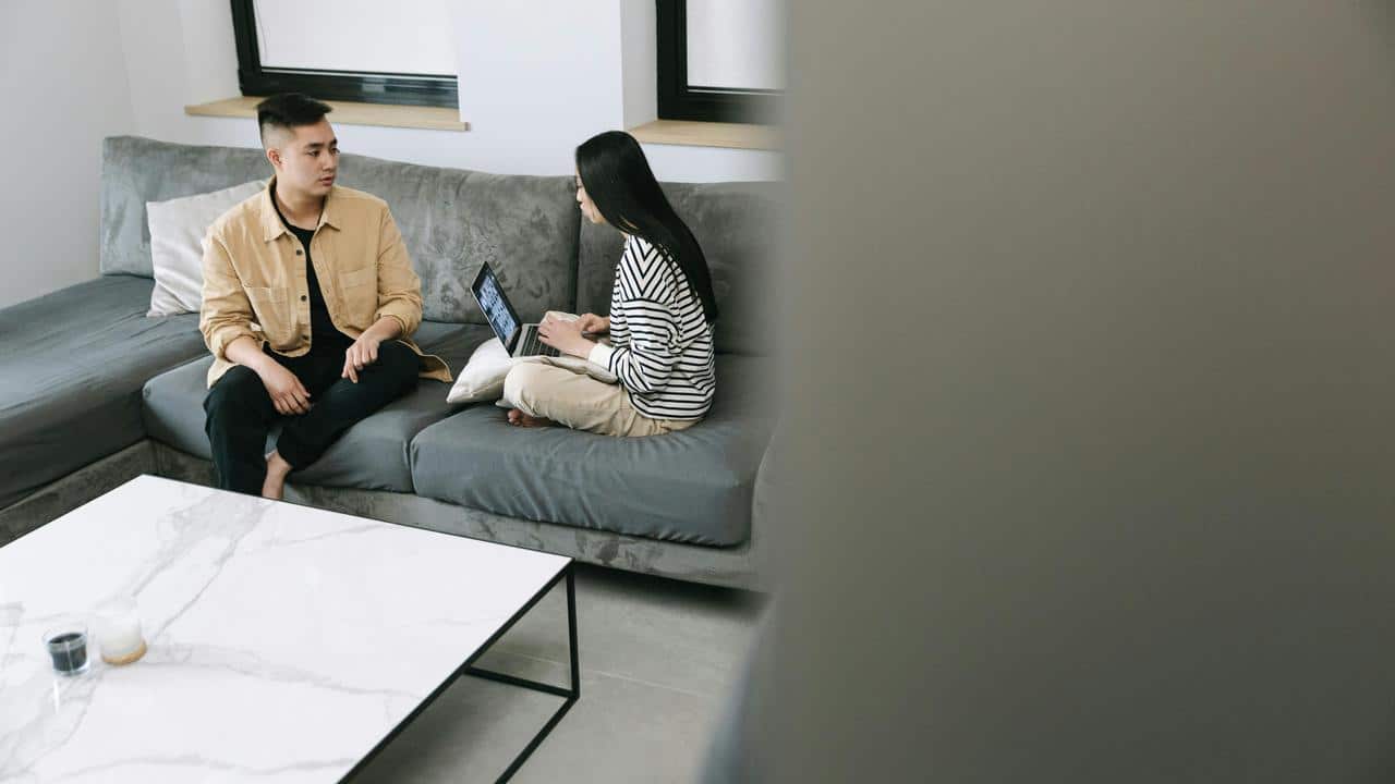 A couple sitting on a couch, talking while using a laptop.
