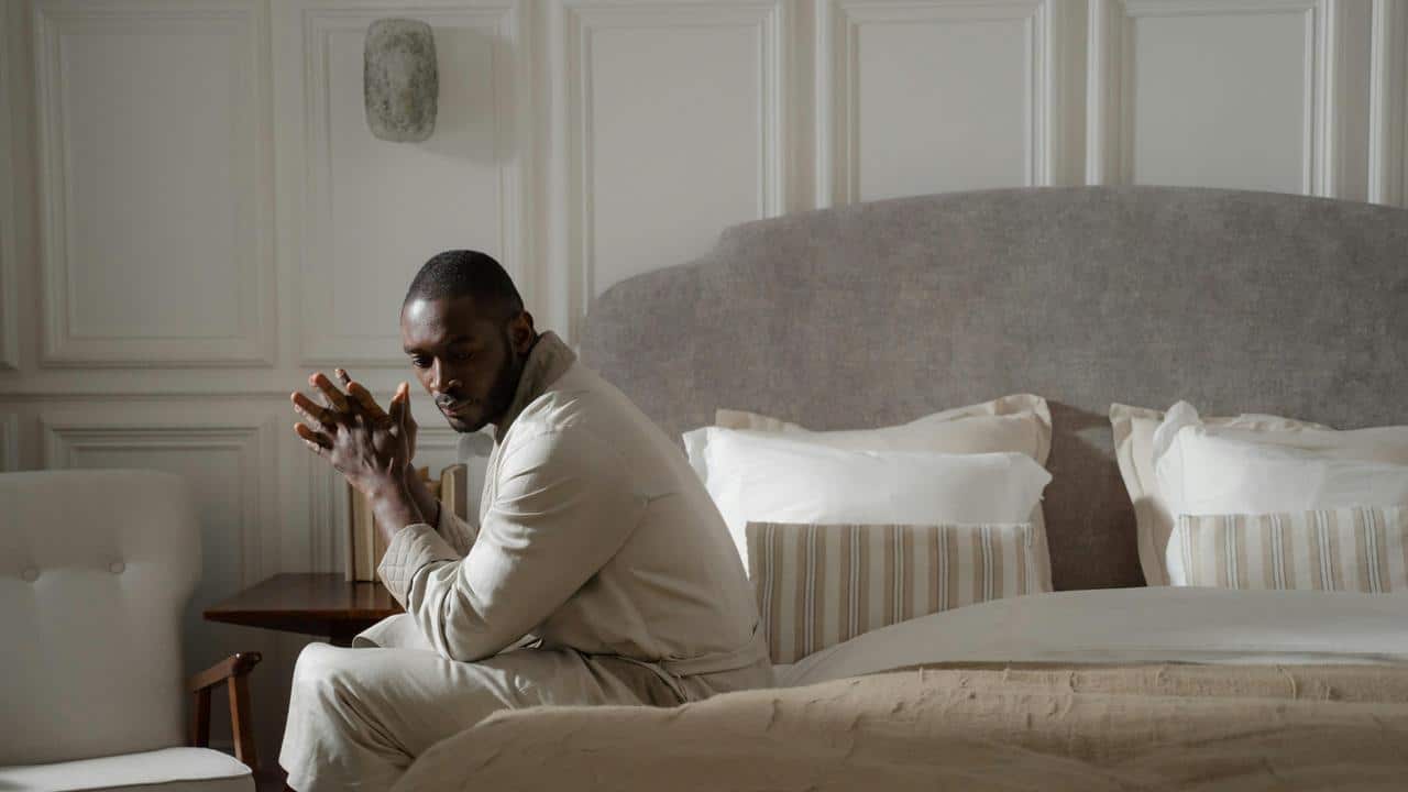 A man sitting on the edge of a bed with his hands clasped, looking thoughtful.