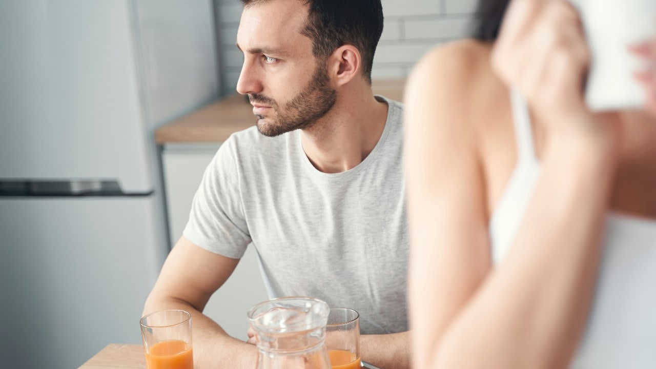 A man sits at a table with orange juice, looking sideways as a woman drinks coffee nearby.