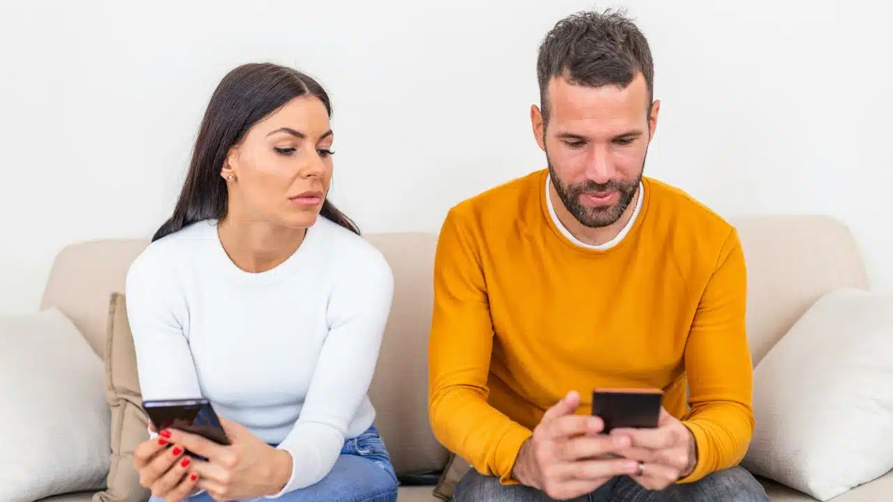 A woman glances sideways at a man while both hold and look at smartphones.