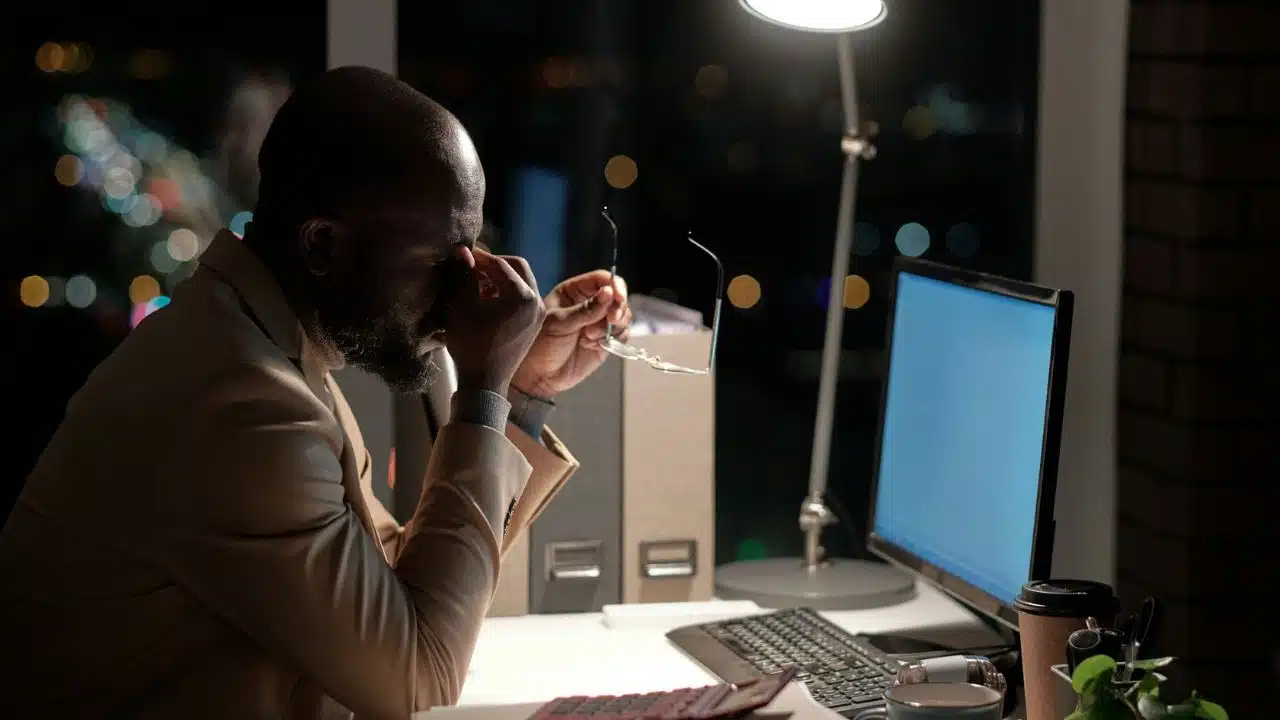 A man rubs his eyes at a desk at night with a computer screen lit.