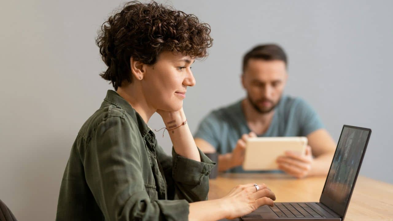 A woman with curly hair uses a laptop while a man uses a tablet behind.