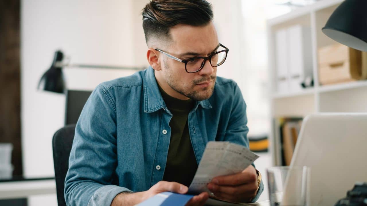 A man with glasses and a denim shirt looks intently at some papers in his hands.