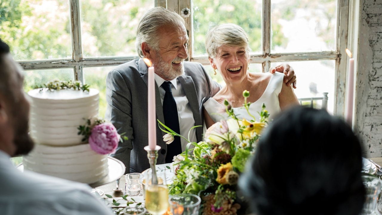 An elderly couple, dressed formally, smiling and laughing at a wedding reception table with a tiered cake and flowers.