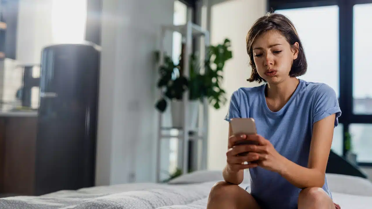 A young woman sitting on a bed looking at her smartphone with a frustrated expression.