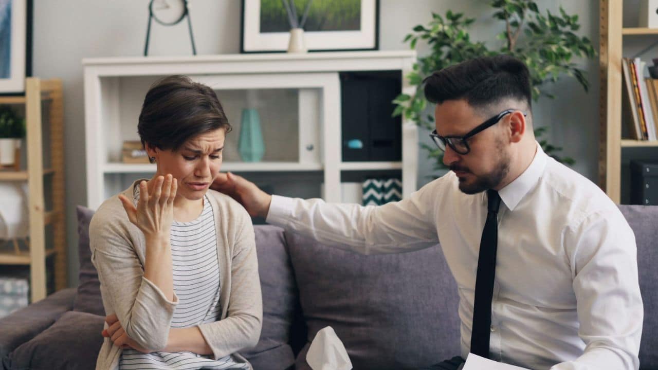 A distressed woman crying, consoled by a man in a white shirt and tie on a couch.