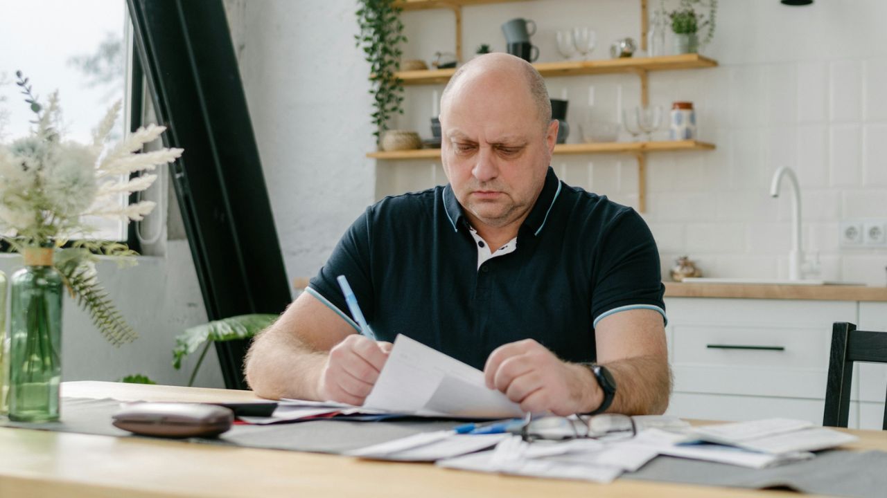 A bald, serious man sitting at a kitchen table reviewing documents and writing.