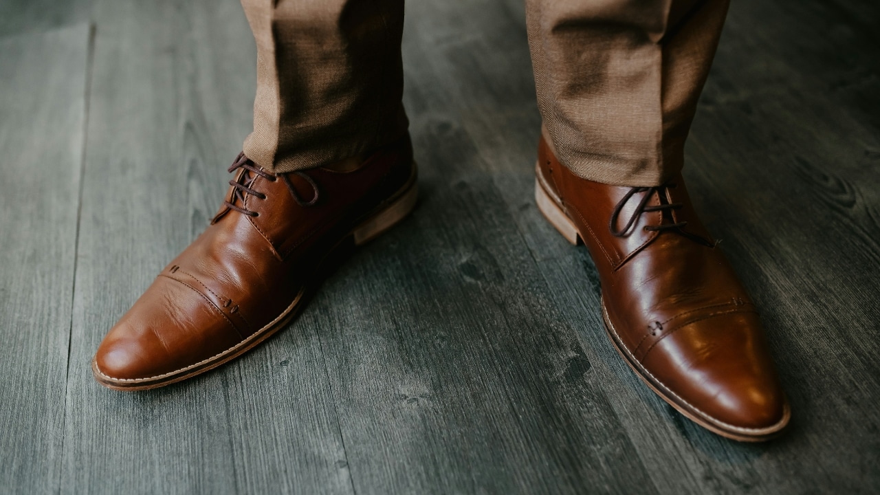 A person wears brown leather dress shoes and tan trousers while standing on wooden flooring.