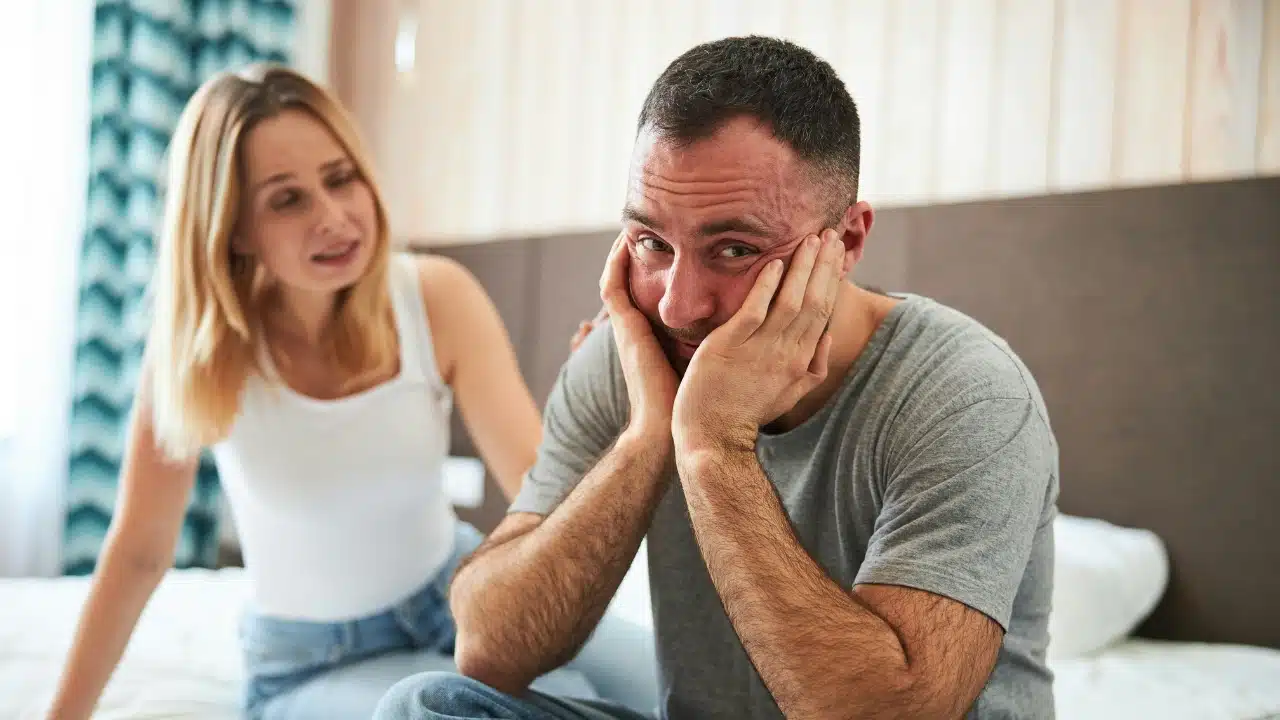 A man sits on a bed with his head in his hands near a woman.