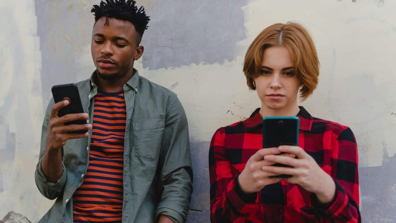 A man and a woman stand against a wall, both looking down at their smartphones.