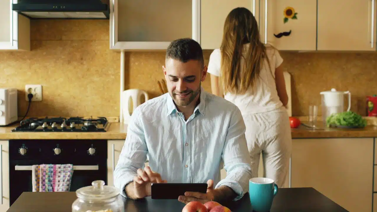A man smiles while using a tablet as a woman works in the kitchen background.