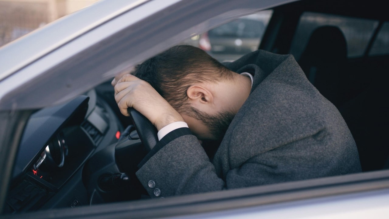 A man in a grey coat rests his head on a car steering wheel, looking down.