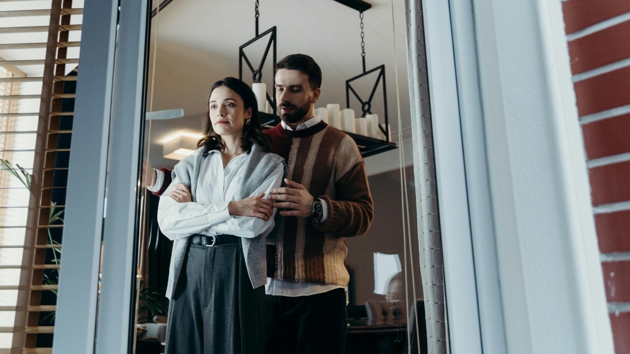 A man stands behind a woman with his hand on her arm near a window.