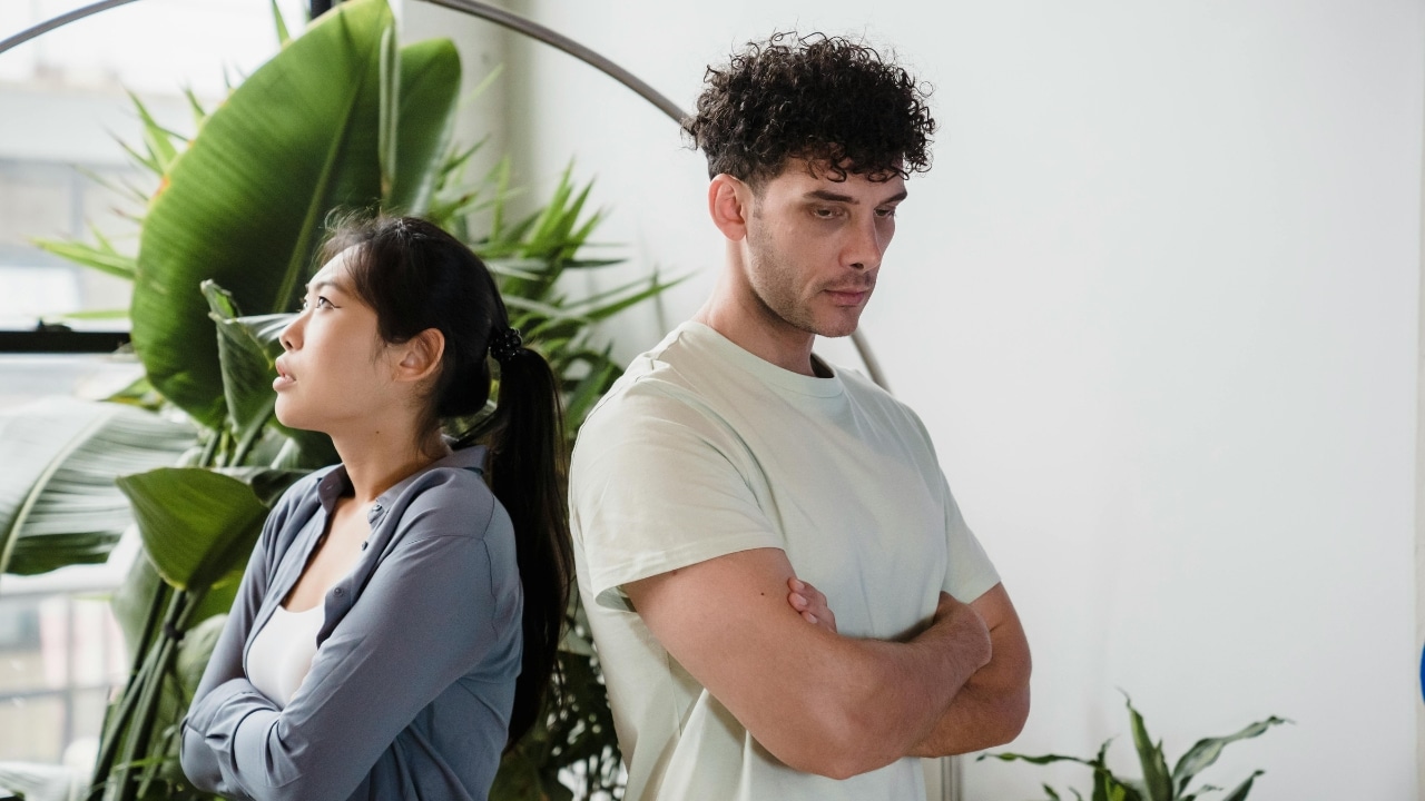 A man and woman stand back to back with their arms crossed in a room.