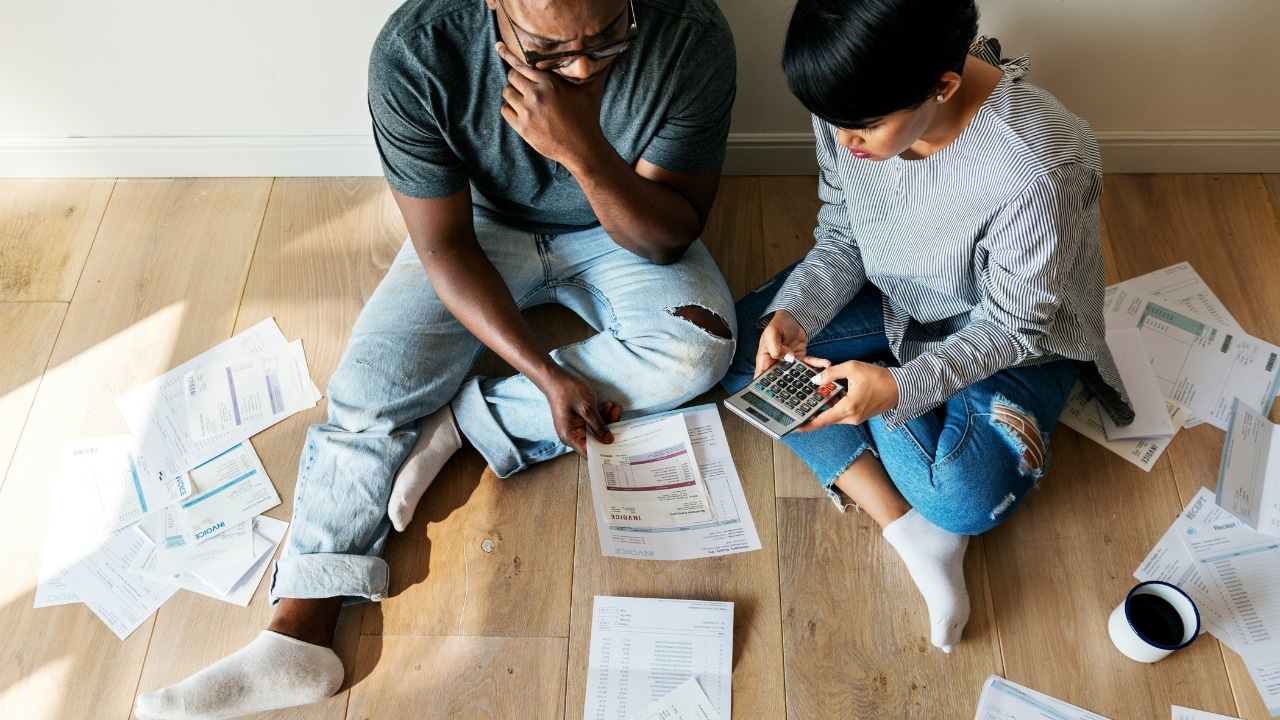 Two people sit on a wooden floor surrounded by many papers and a calculator.