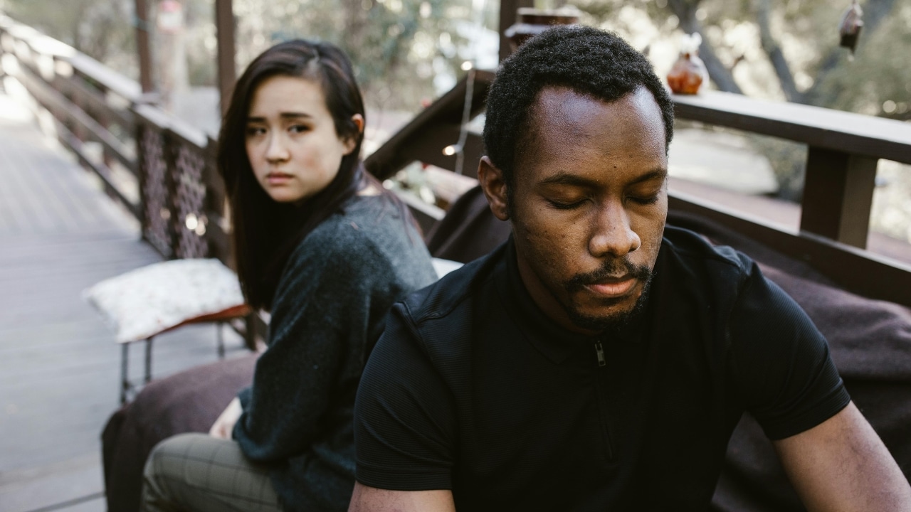 A man with eyes closed sits in the foreground with a woman looking at him.