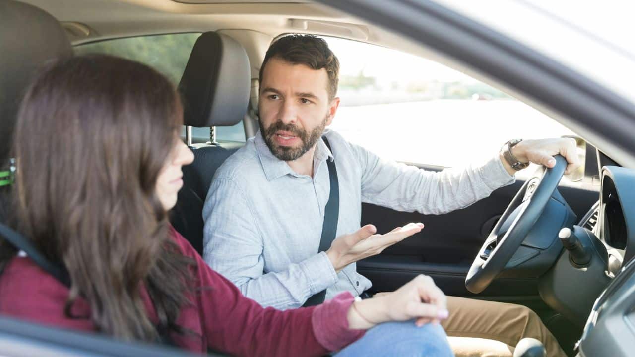 A bearded man talks while driving a car with a woman sitting in the passenger seat.