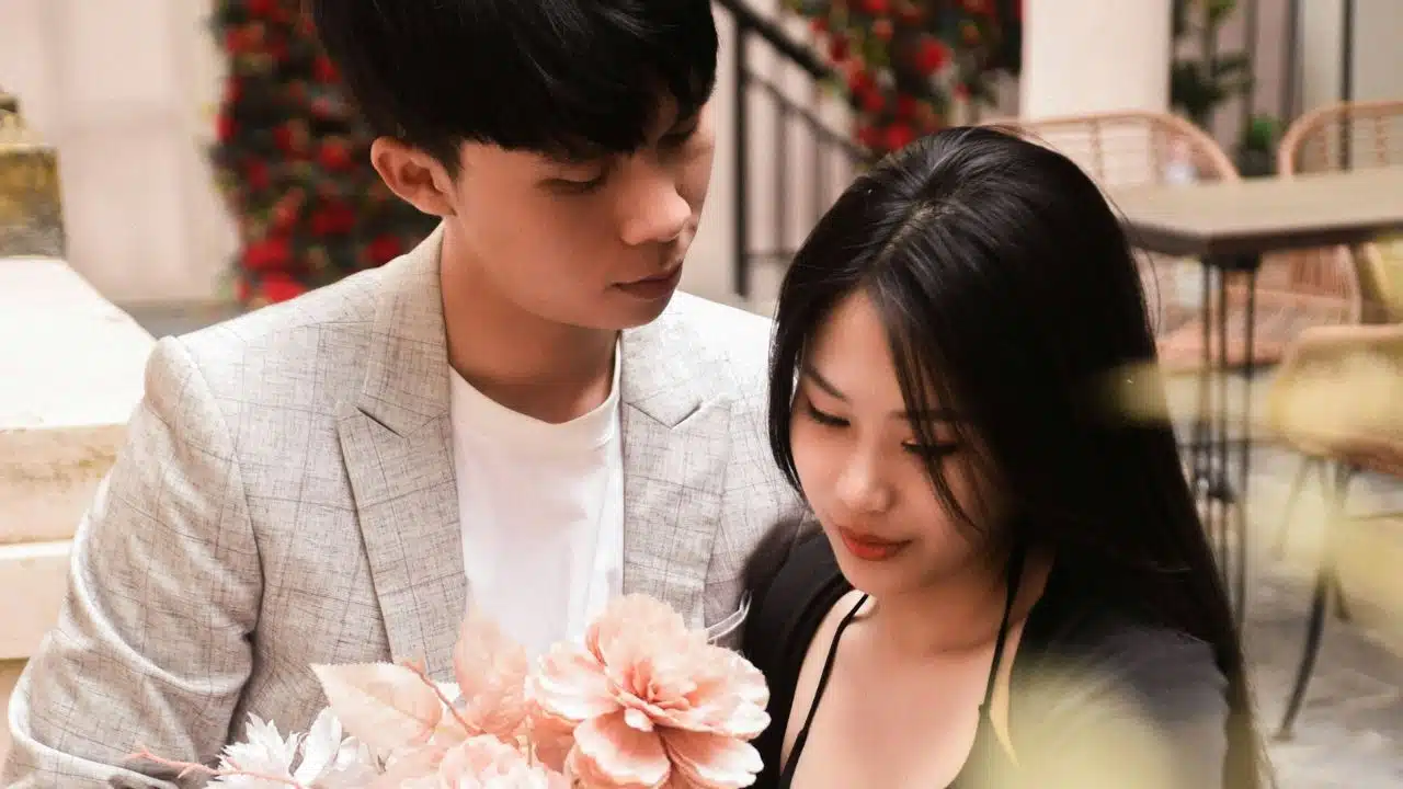 A young man and woman look down together at a bouquet of large pink flowers.