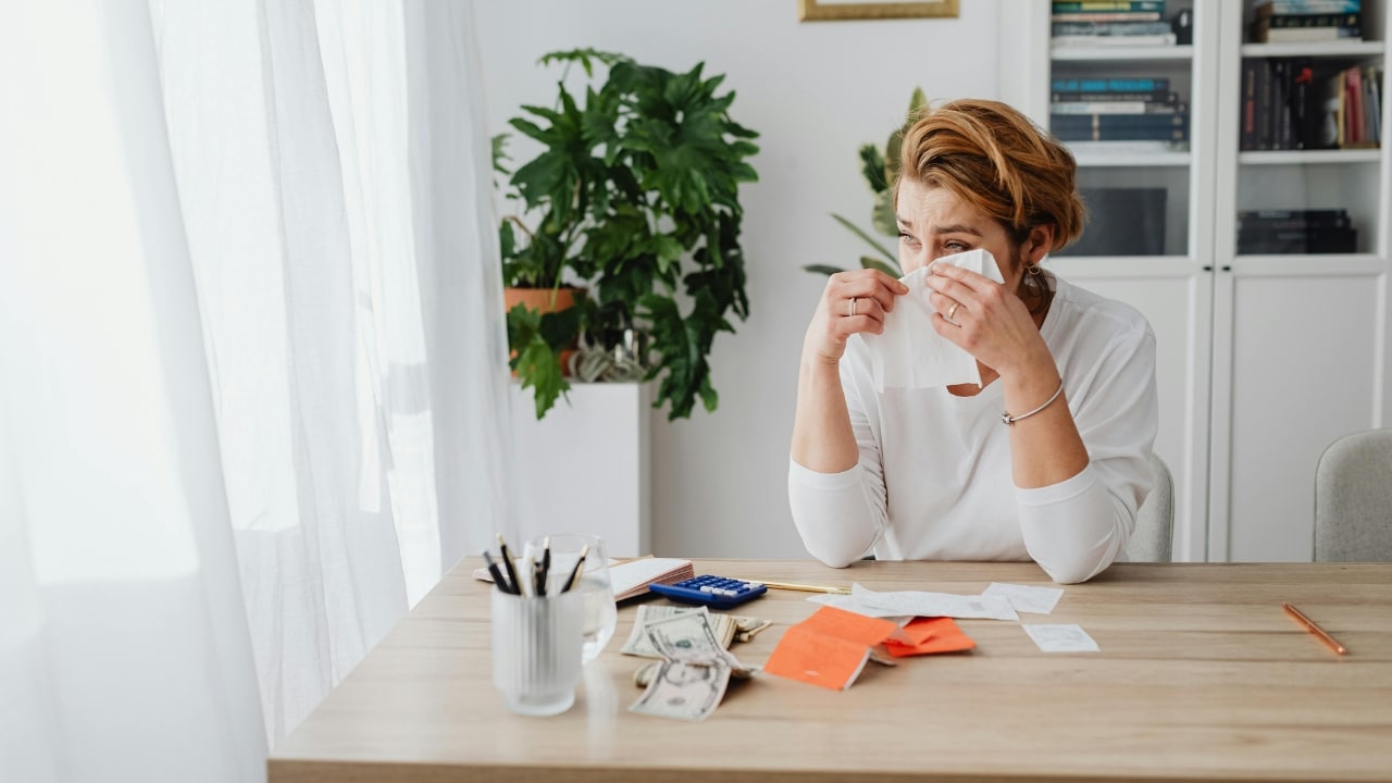 A woman sits at a desk with papers and money, holding a tissue to her face.