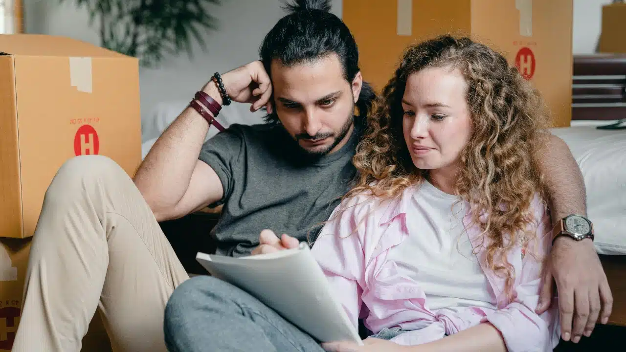 A man and woman sit on the floor among cardboard boxes looking at a notebook.