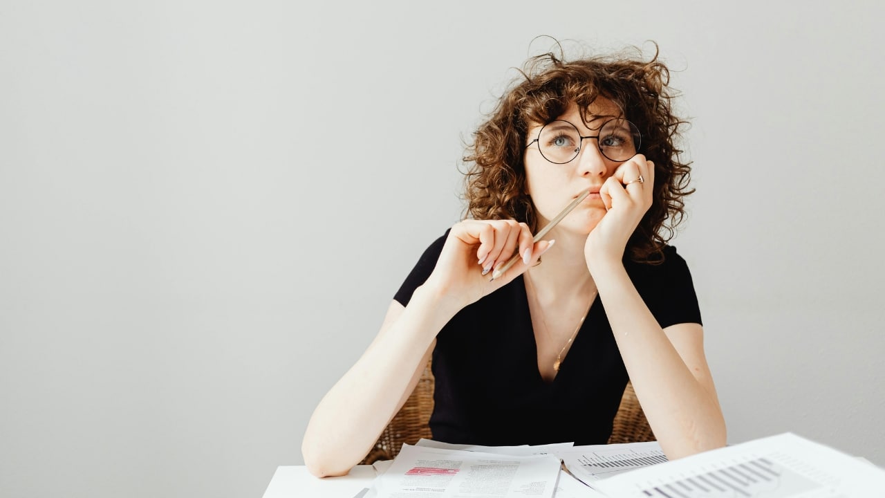 A woman with glasses holds a pencil to her face while looking upward at papers.