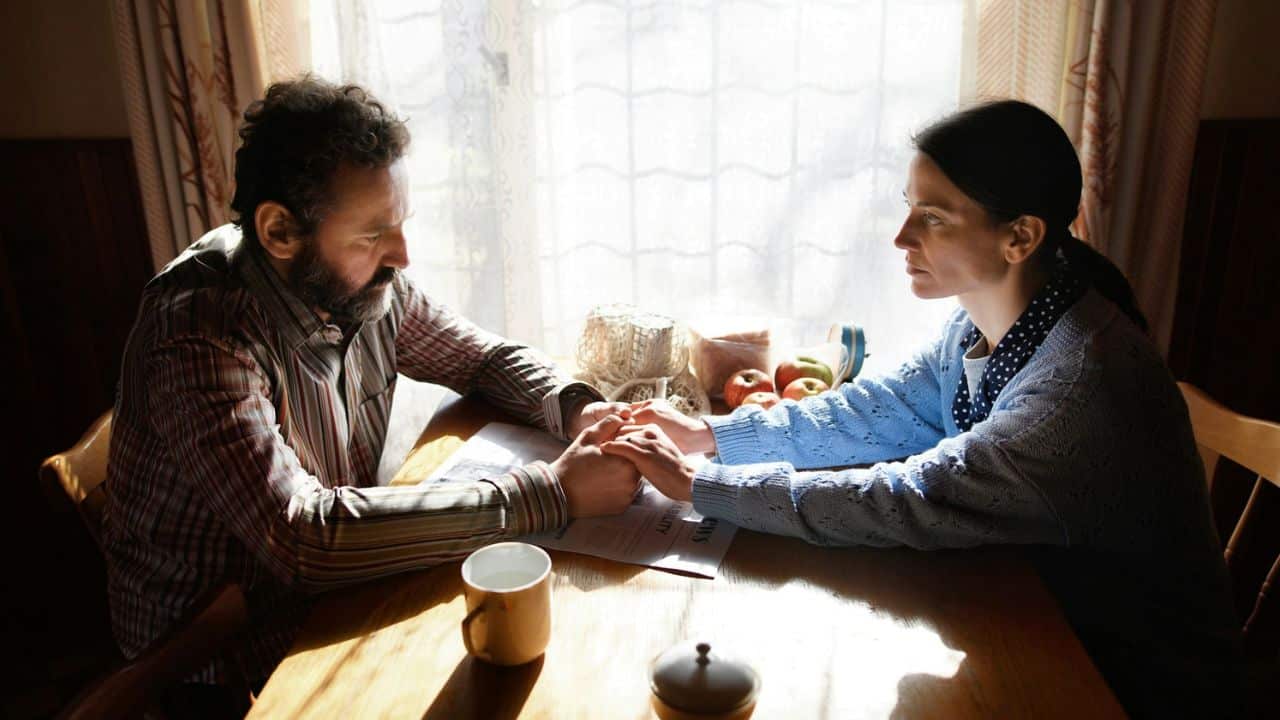 A man and woman sit at a wooden table holding hands in bright sunlight.
