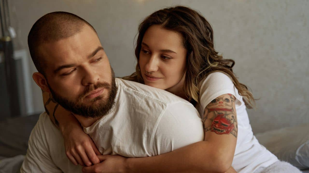 A woman with a tattoo on her arm hugs a bearded man from behind.