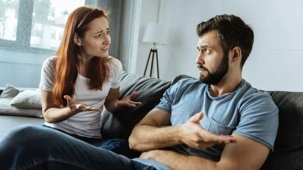 A woman with a distressed expression gestures toward a man who looks away while gesturing.