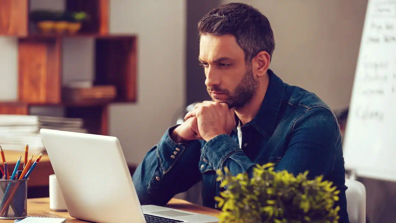 A man in a denim shirt sits at a desk, looking intently at a laptop screen.