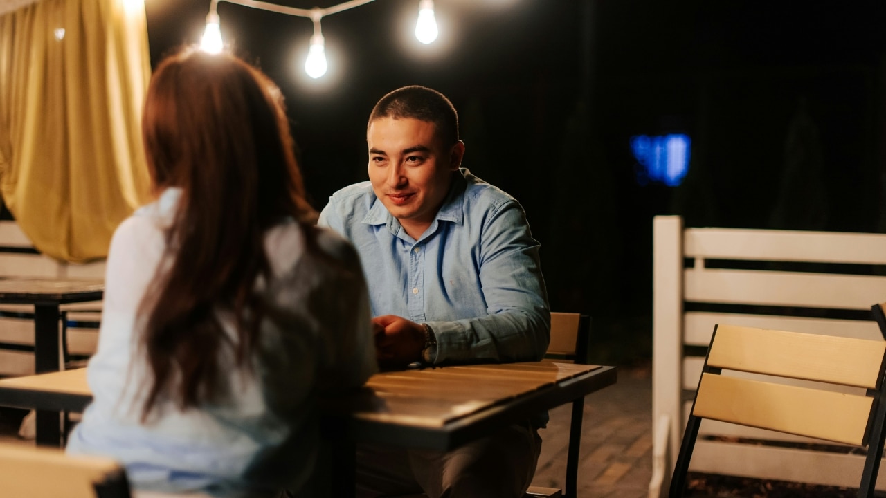 A man in a blue shirt sits at an outdoor table at night, looking at a woman.