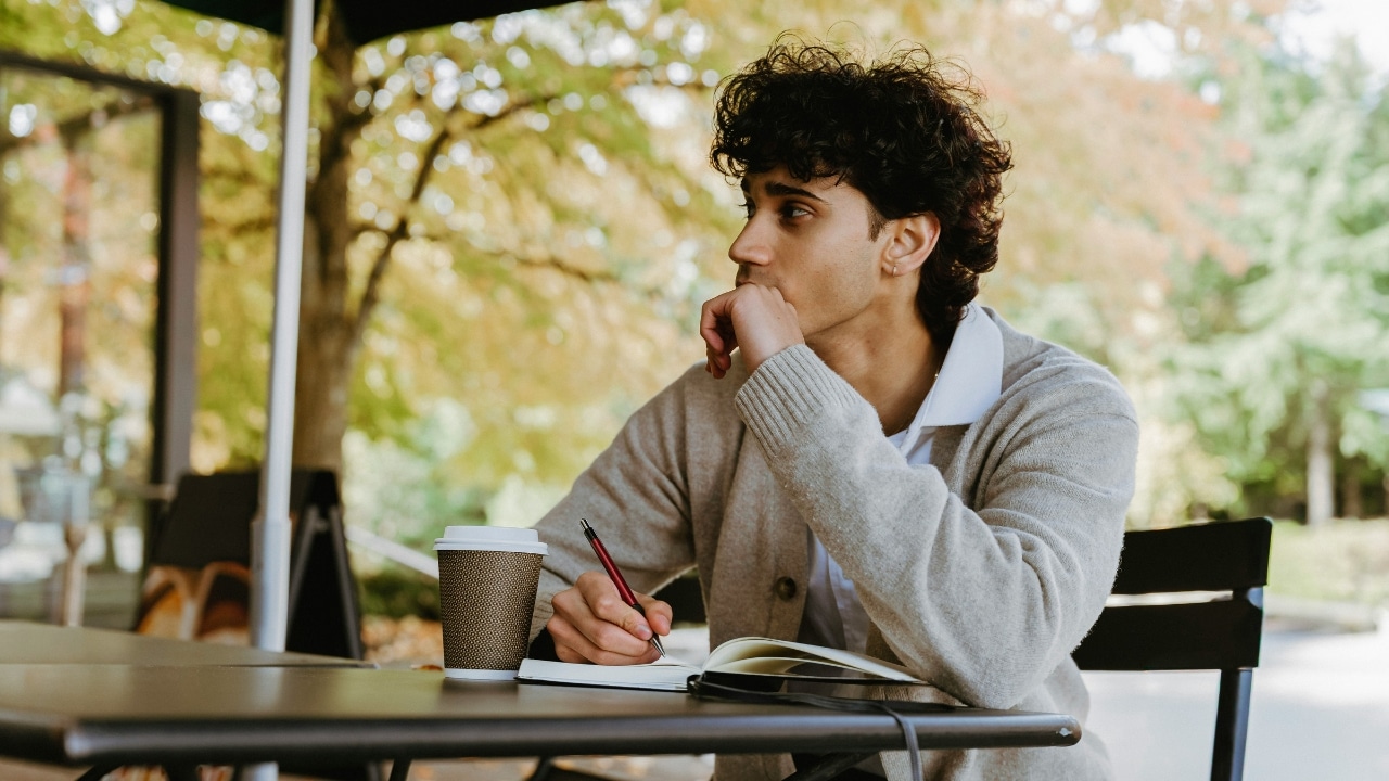 A young man with curly hair sits at an outdoor table writing in a notebook.