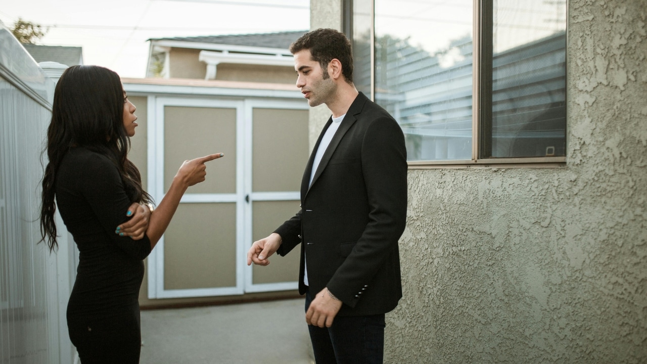 A woman in a black dress points her finger at a man wearing a black blazer outdoors.