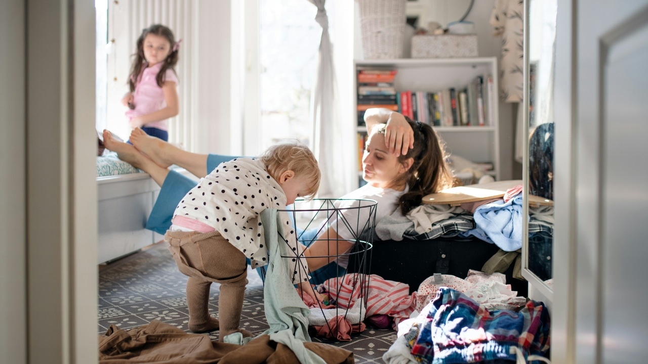 A woman sits on the floor near a laundry basket while a child reaches inside.