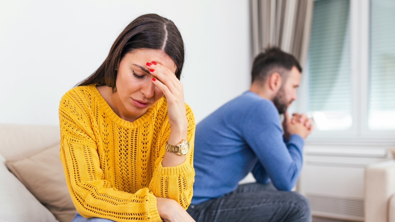 A woman in a yellow sweater touches her forehead while a man sits behind her.