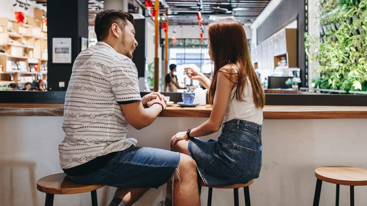 A man and woman sit on stools at a wooden counter talking in a cafe.